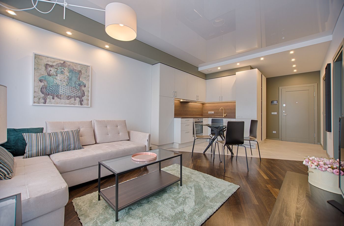 A living room lit by recessed ceiling downlights and a central pendant, with a sofa and dining table beyond