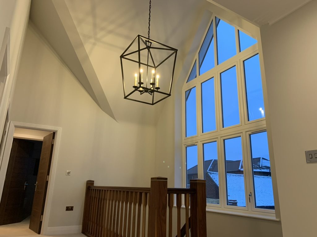 A black lantern pendant in a double-height room with a gable window — drop height set so it reads from both the ground floor and the landing.